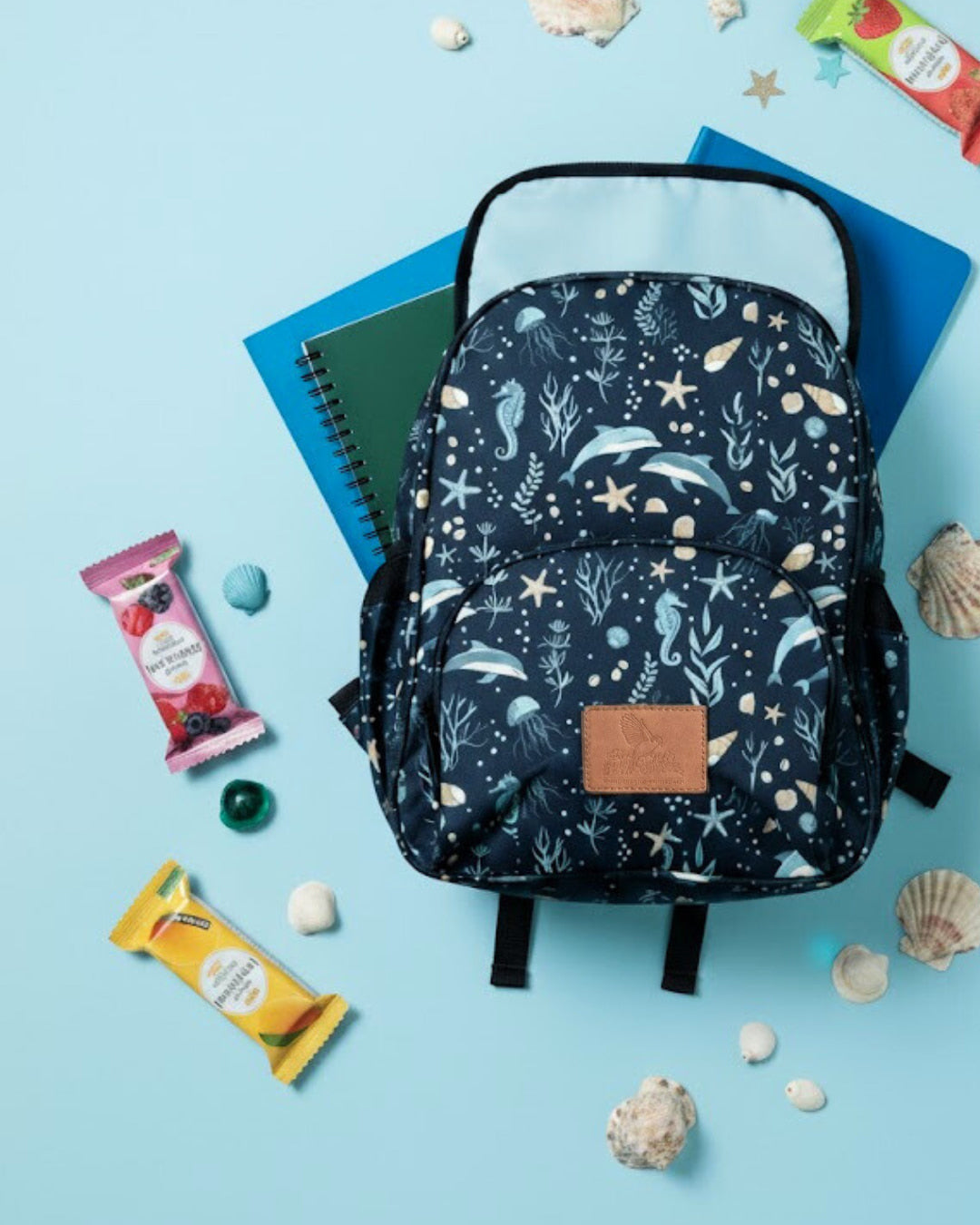 Navy blue backpack with sea-themed pattern on a light blue background with snacks and shells.