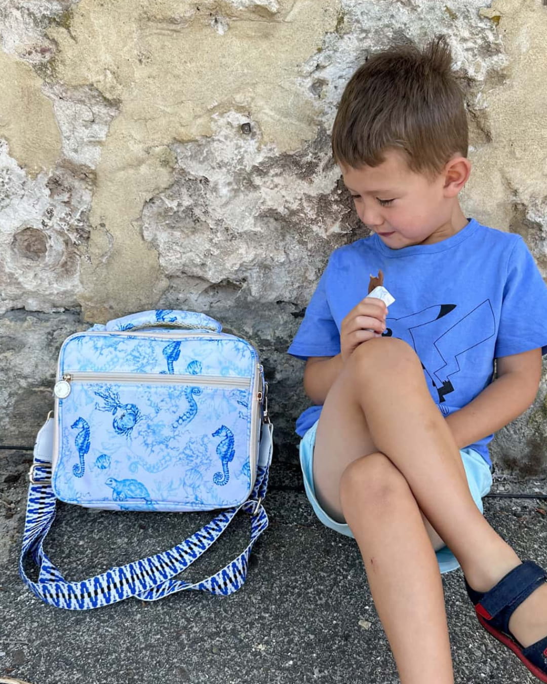 Boy sitting next to blue ocean print insulated lunch bag with stylish cross-body strap for kids' school and day trips.