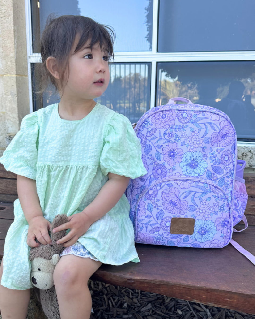 Child in a light green dress sitting next to a purple backpack with floral patterns on a wooden bench.