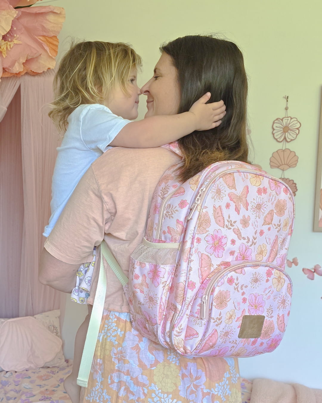 Mum wearing pink butterfly print small backpack as a stylish nappy bag while holding her child for family outing.