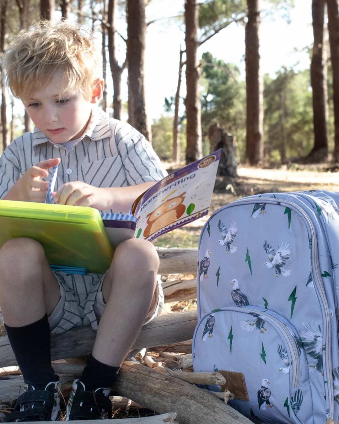 Boy sitting outdoors with light blue eagle print small backpack for school, handwriting practice, and forest trips.