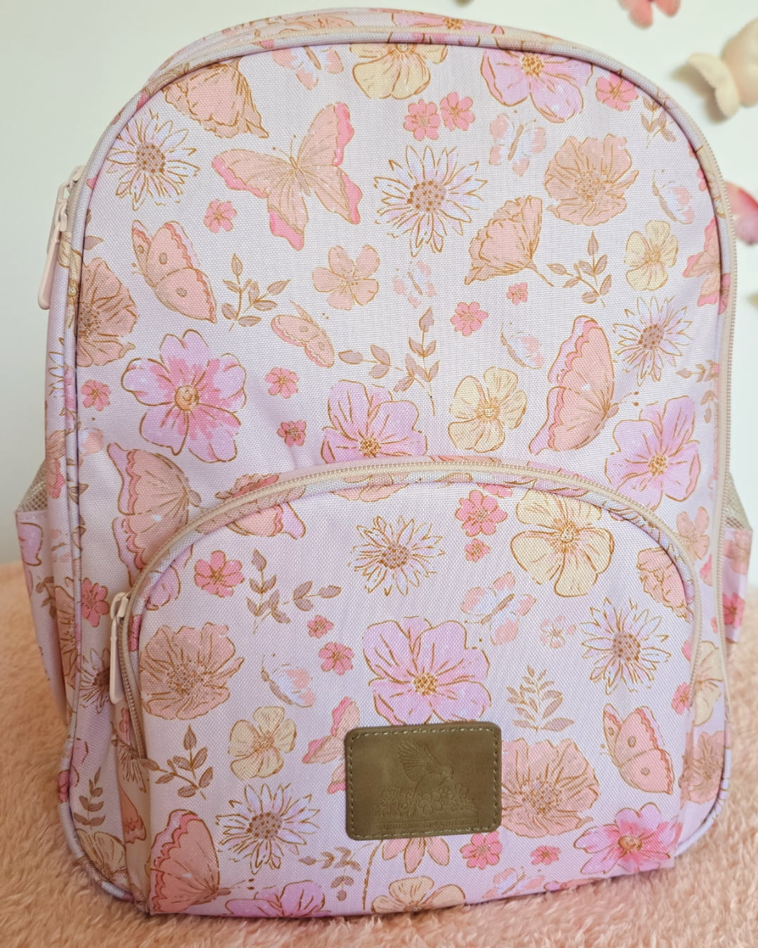 Close-up of pink butterfly and floral print small backpack showing front pocket and leather-look brand patch.