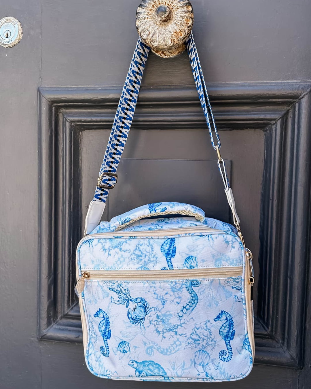 Insulated lunch bag with blue ocean print and adjustable cross-body strap, hung on door handle for easy travel.