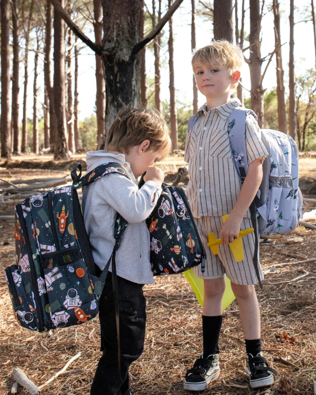 Two boys outdoors wearing small backpacks in space theme and light blue eagle print for siblings and family adventures.