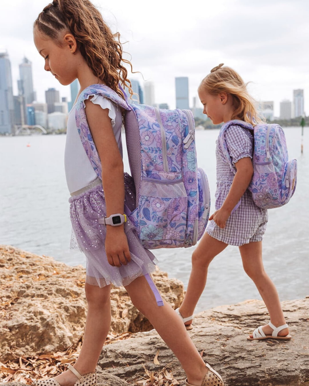 Two girls walking by city skyline with lilac floral small backpacks, showing size and fit for children's school or travel bags.