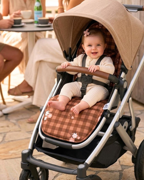 Baby in a stroller with a checkered pad, sitting outdoors with people in the background.