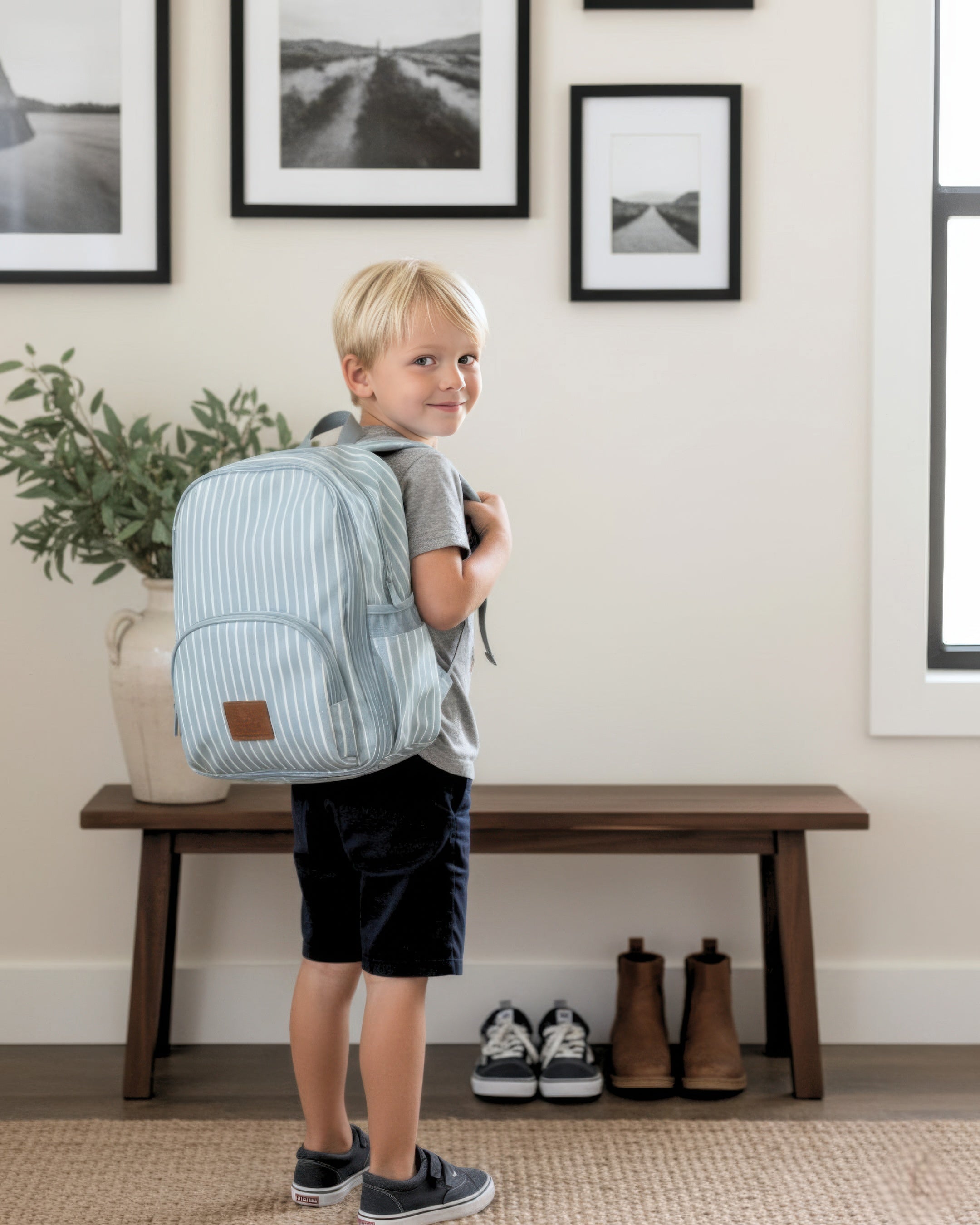 Child with a striped backpack standing in a room with framed pictures on the wall.