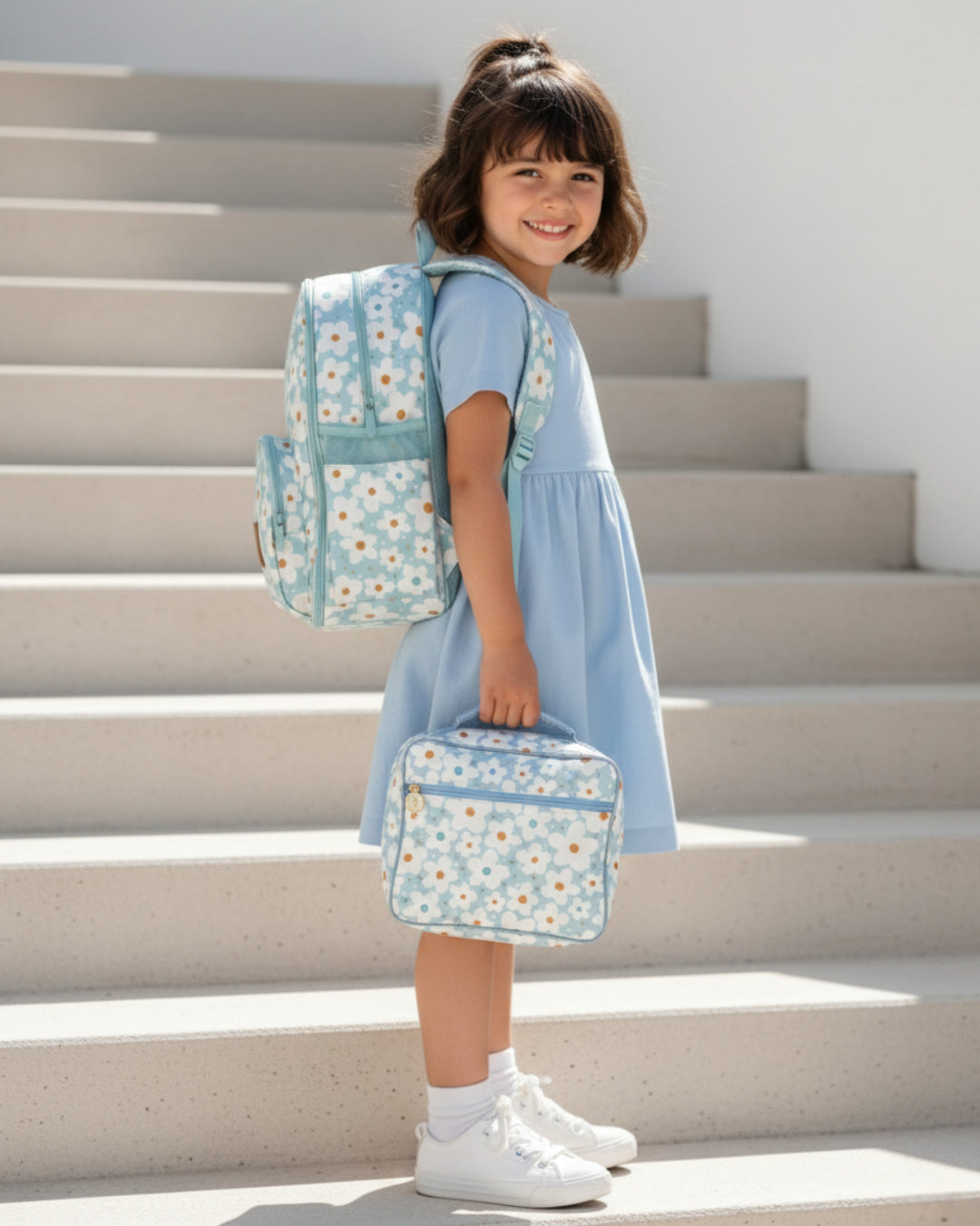 Child in a blue dress with floral backpack and suitcase on steps