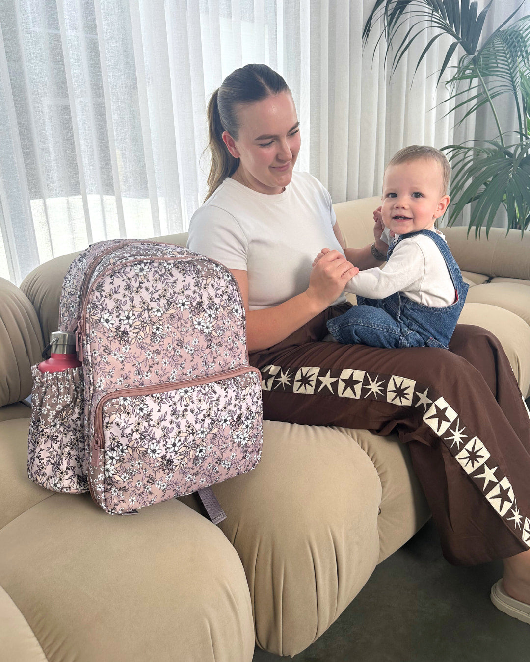 Woman sitting on a couch with a child, next to a pink backpack with floral pattern.