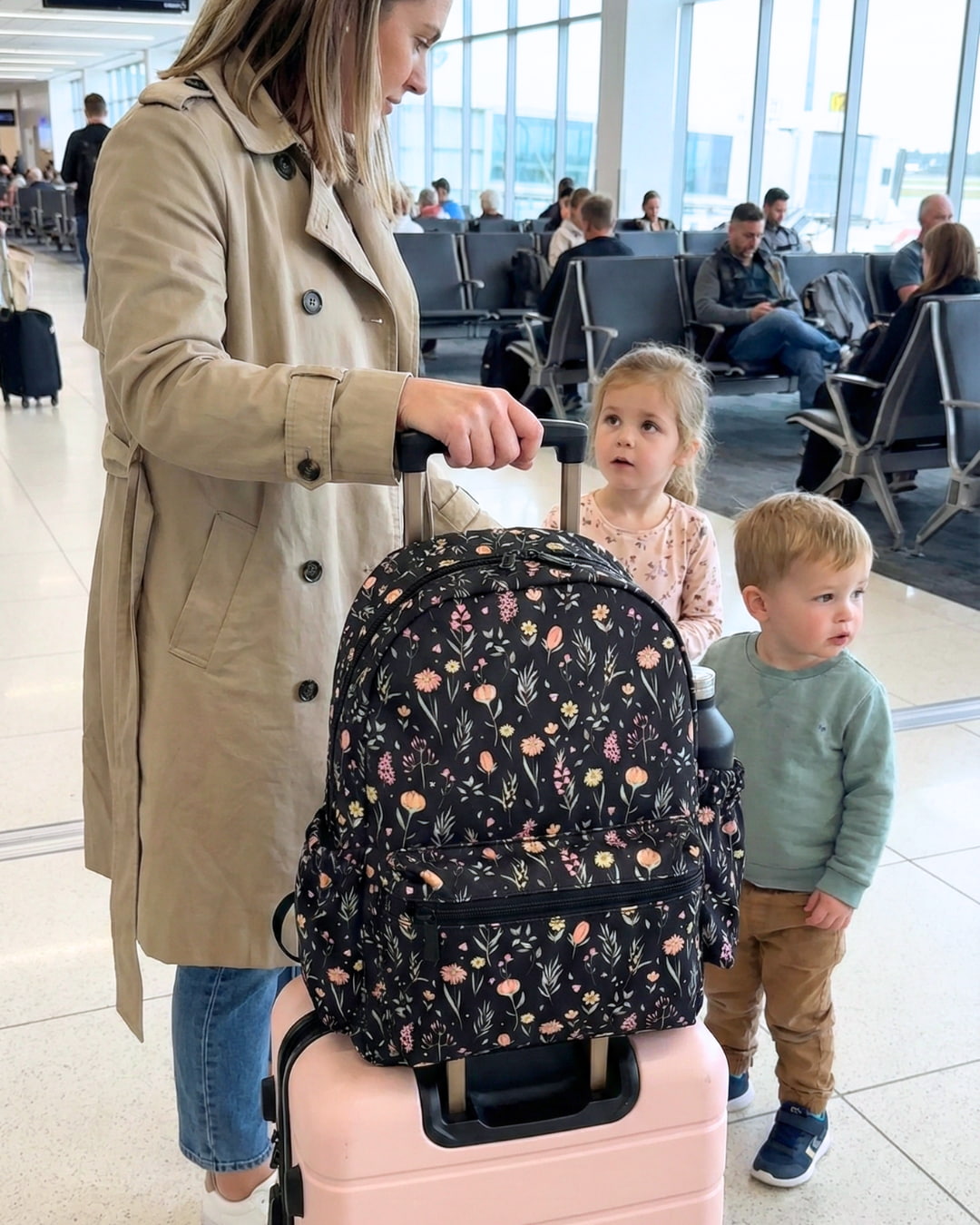 Woman with a floral backpack and pink suitcase in an airport with two children.
