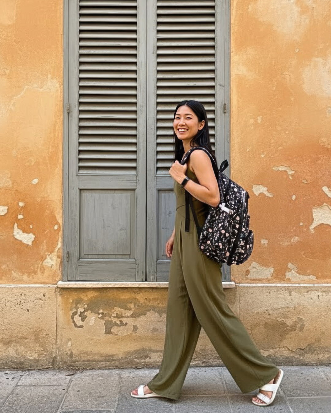 Woman in a green jumpsuit with a black floral backpack standing in front of a textured wall.