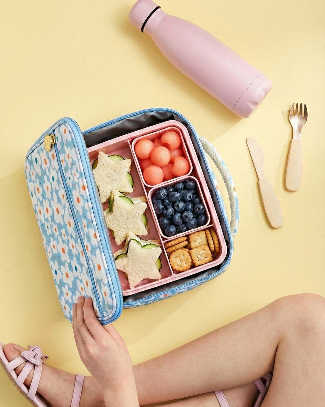 Lunchbox with compartments filled with food, a pink water bottle, and utensils on a yellow background.