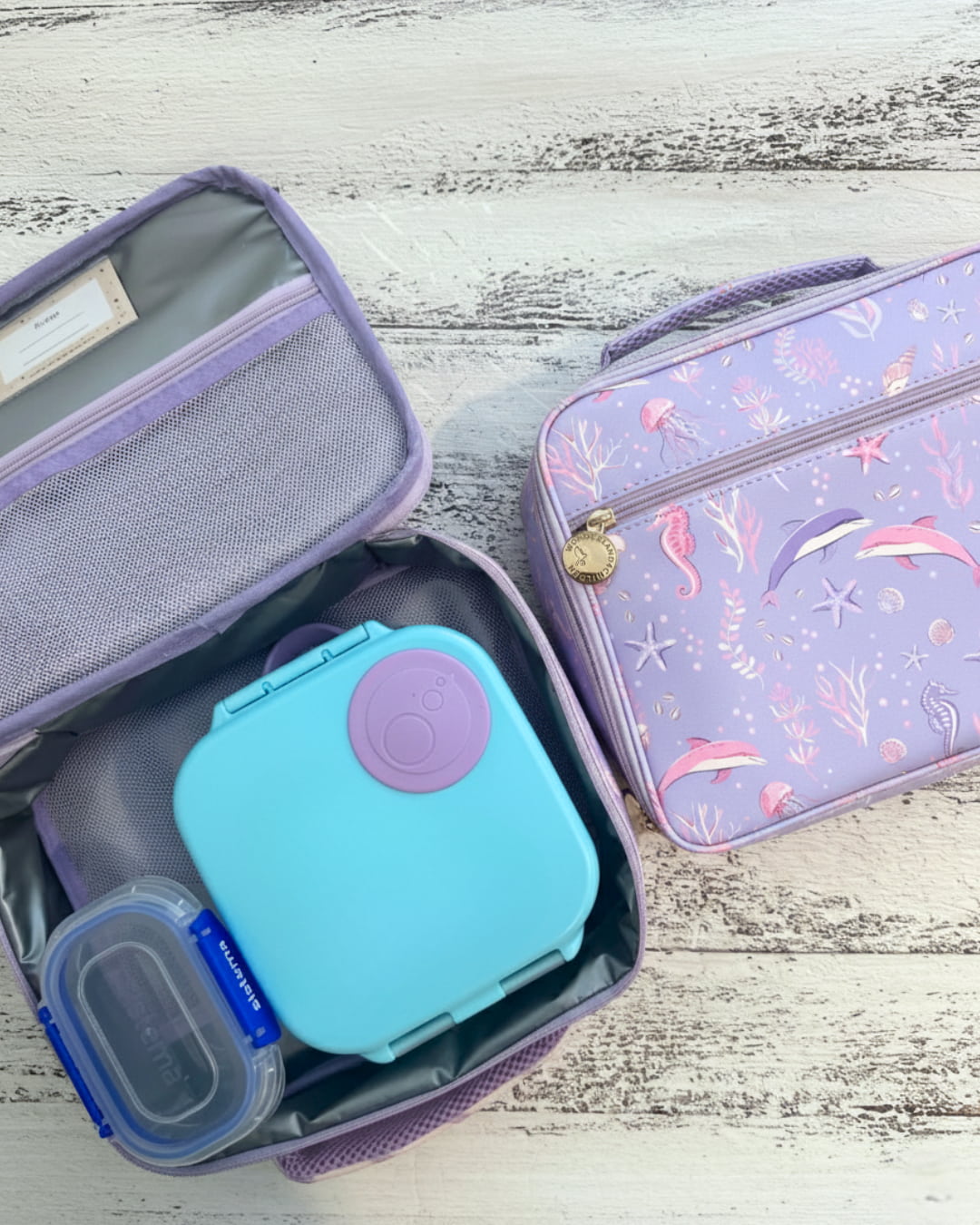 Purple lunch bag with compartments containing a teal container and other items on a wooden surface