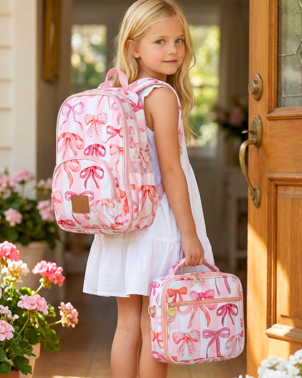 Young girl with a pink backpack and matching lunch bag in front of a wooden door.