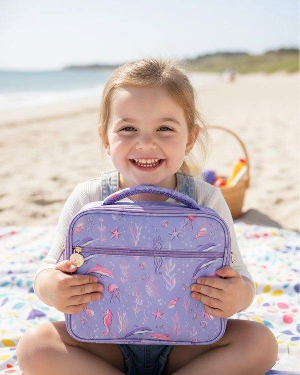 Child holding a purple lunch bag with mermaid design on a beach