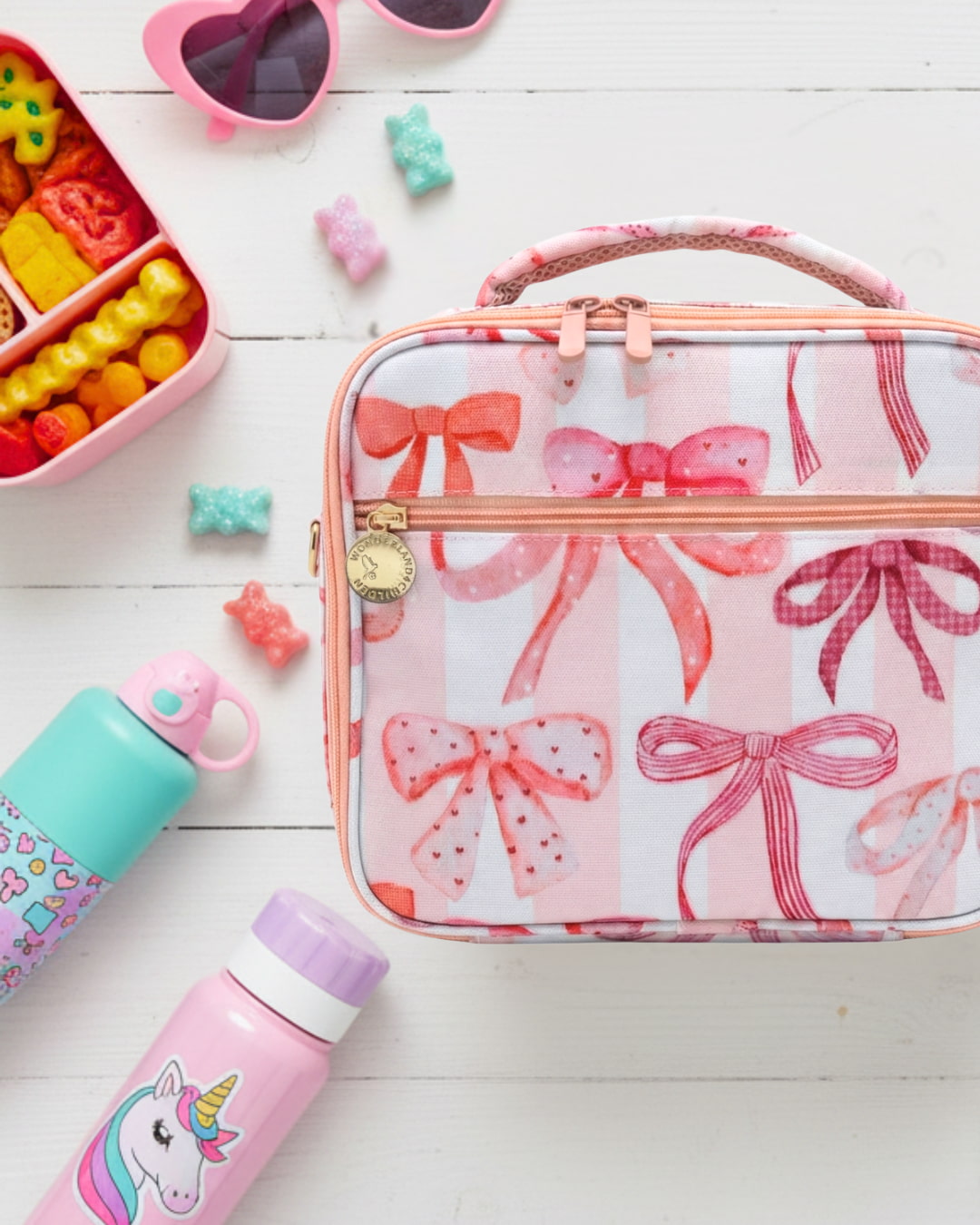Pink lunch bag with bow pattern, colorful water bottles, and snacks on a white surface