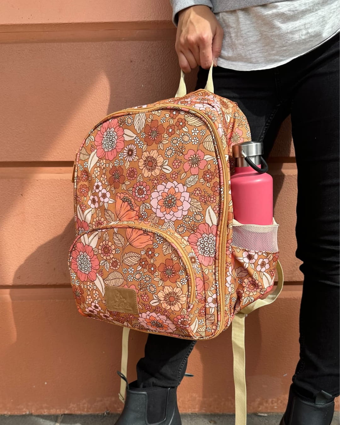 Person holding a floral-patterned backpack with a pink water bottle against a wall.