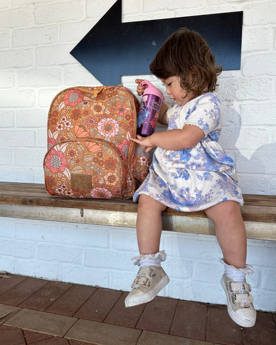 Child sitting on a bench with a floral backpack and pink water bottle.