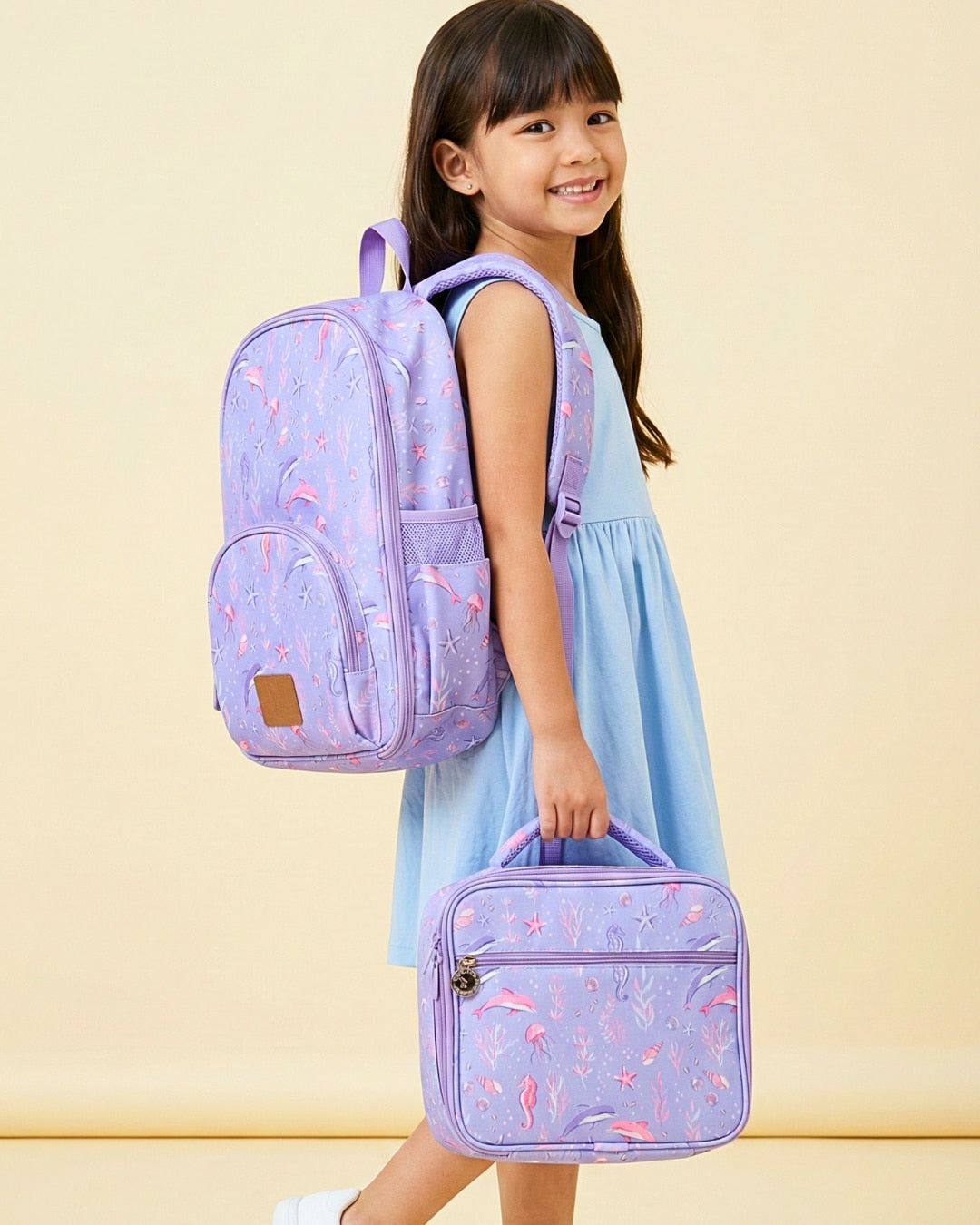 Young girl wearing a purple backpack and holding a matching lunch bag against a beige background