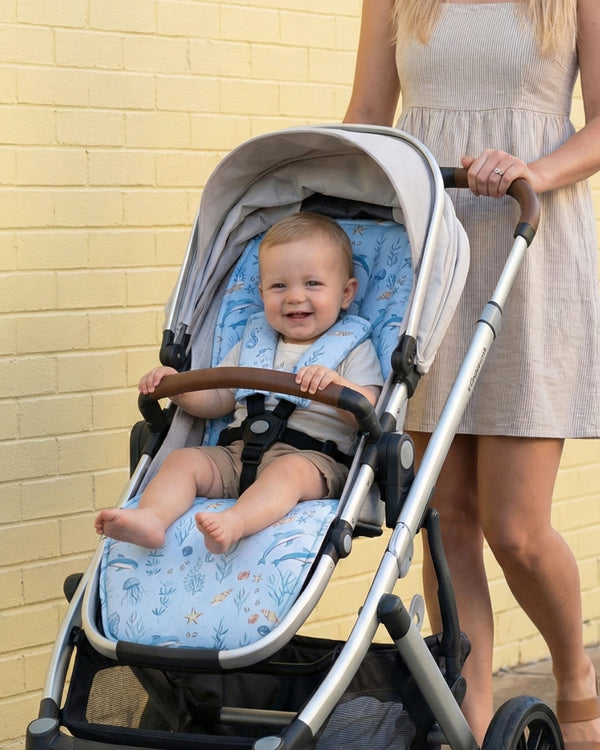 Baby in a stroller with a woman standing behind it against a beige brick wall.