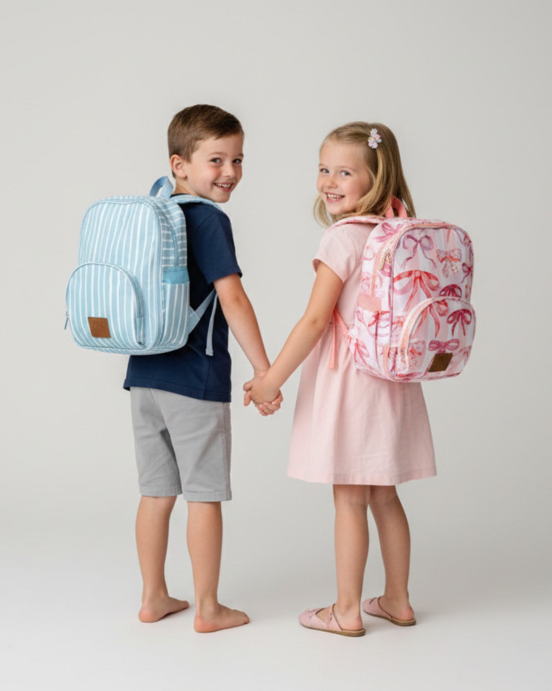 Two children holding hands, each wearing a colorful backpack on a plain background