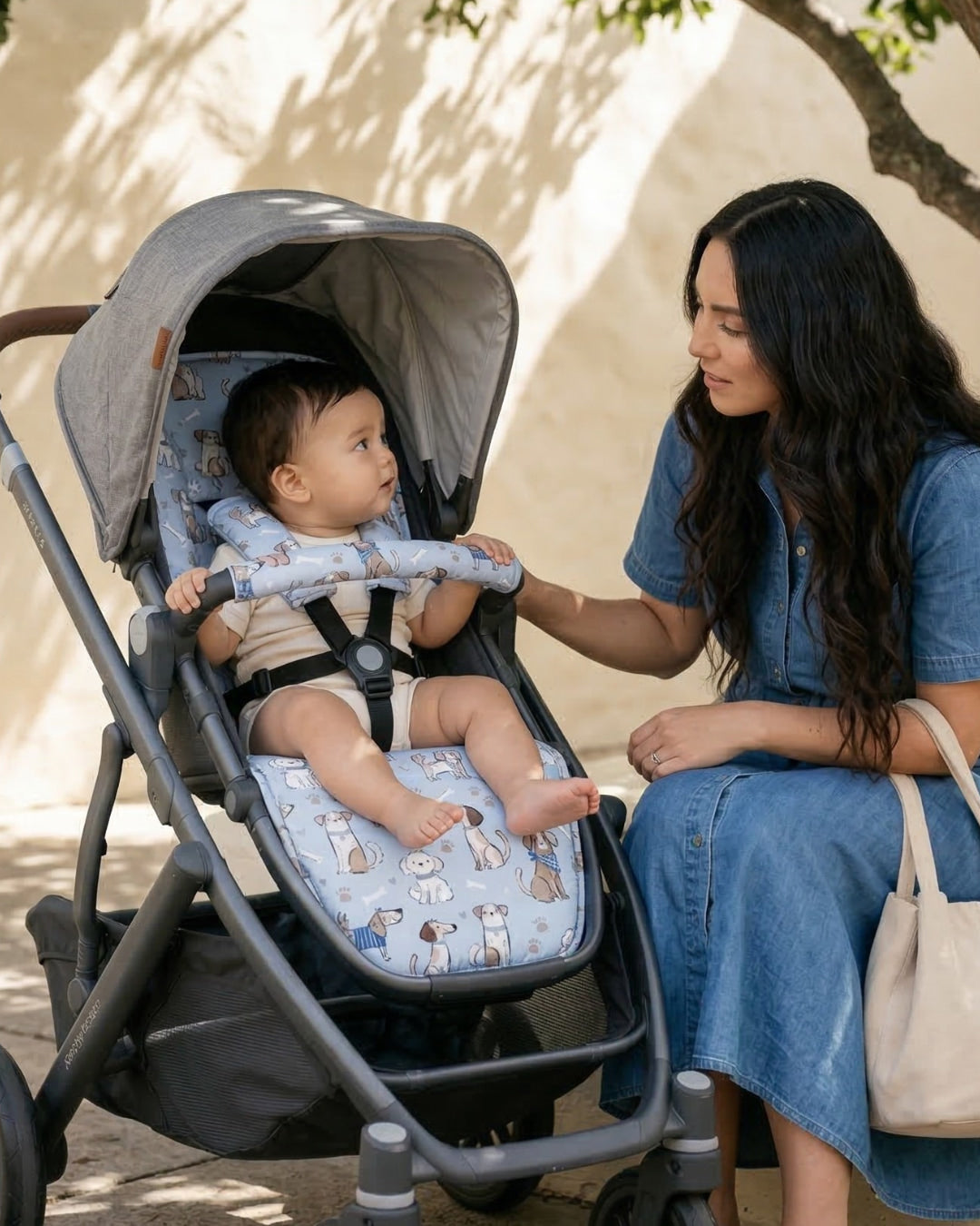 Woman sitting beside a stroller, gently touching a baby seated inside on a light blue dog-patterned stroller liner while they look at each other outdoors in soft shade.