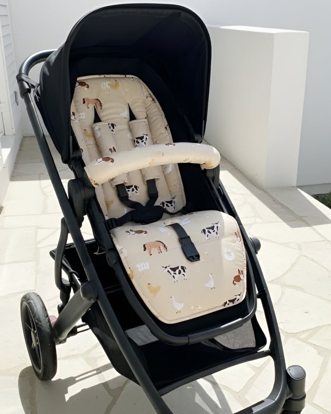 Empty stroller fitted with a beige farm animal-print pram liner and matching padded bumper bar cover, displayed outdoors in sunlight.