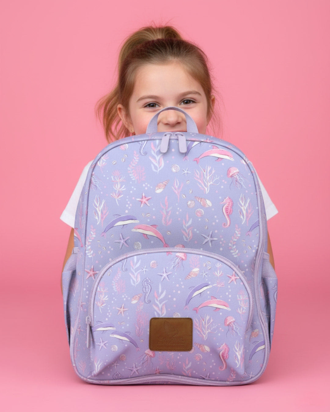 Child holding a purple backpack with mermaid pattern against a pink background