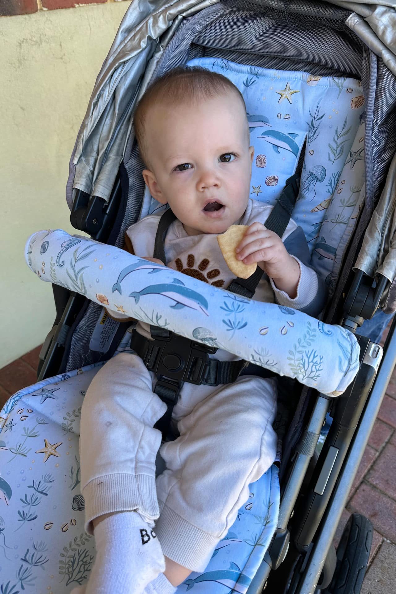 Baby sitting in a stroller with a patterned ocean themed seat cover, holding a snack.