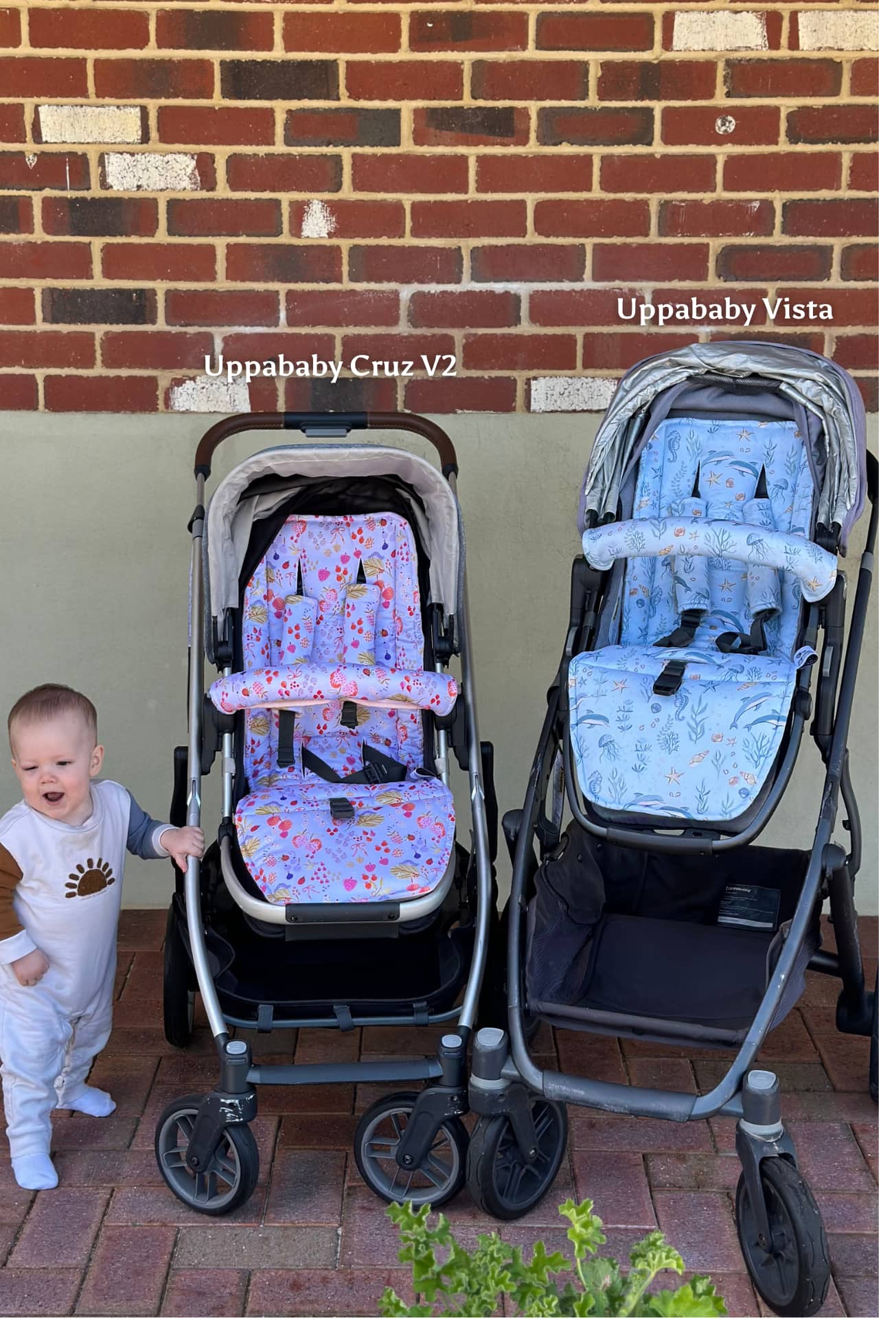 Two strollers, one Uppababy Cruz V2 and one Uppababy Vista, with a child standing next to them against a brick wall.
